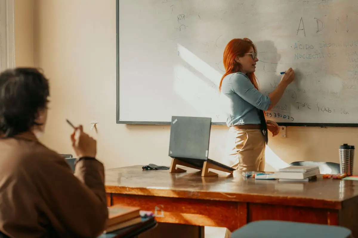 Docente haciendo clases frente a la pizarra, con su notebook abierto sobre el escritorio.